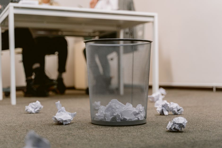 A grey metal mesh waste bin situated on a beige carpeted office floor, partially filled with crumpled white paper. Several additional crumpled papers are scattered around the base of the bin, some par