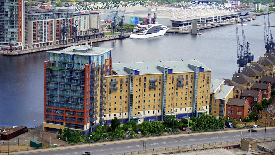 A daytime view of a narrow canal with calm, reflective water running between two modern residential buildings constructed from light brown and beige bricks, with black metal balconies and window frame