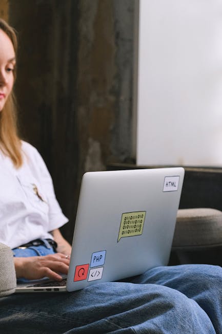 The image shows a young woman with light brown hair sitting indoors, working on a silver laptop placed on her lap. Her face is partially visible on the left side of the frame. The laptop's exterior fe