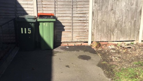 The image shows two waste collection bins placed on a concrete driveway adjacent to a wooden fence. One bin is dark green with the number 115 painted in yellow on its front, and the other bin is also green with a black lid. The bins are positioned side by side, with the darker green one slightly in front. Shadows cast by the fence and bins fall across the driveway, which has some dark patches likely from previous spills or moisture. To the right, part of a weathered wooden gate or fence panel is visible, along with a small patch of bare soil and a few weeds at its base. The scene appears to depict a typical residential rubbish disposal area, relevant to rubbish removal services in London, with the bins suggesting the collection of general waste or recyclables. The overall setting is quiet and utilitarian, with a focus on the practical placement of waste containers on a typical urban property.